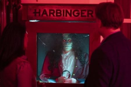 Two people stand in front of an animatronic fortune teller labeled “Harbinger” with glowing red eyes, in a dim, red-lit room.