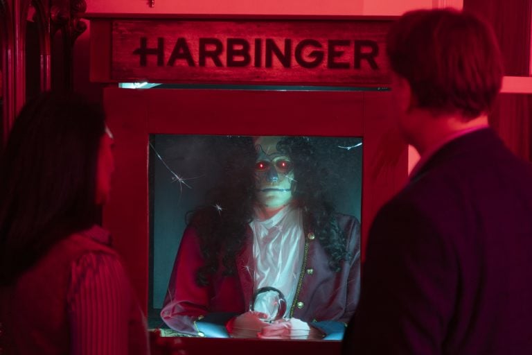 Two people stand in front of an animatronic fortune teller labeled “Harbinger” with glowing red eyes, in a dim, red-lit room.
