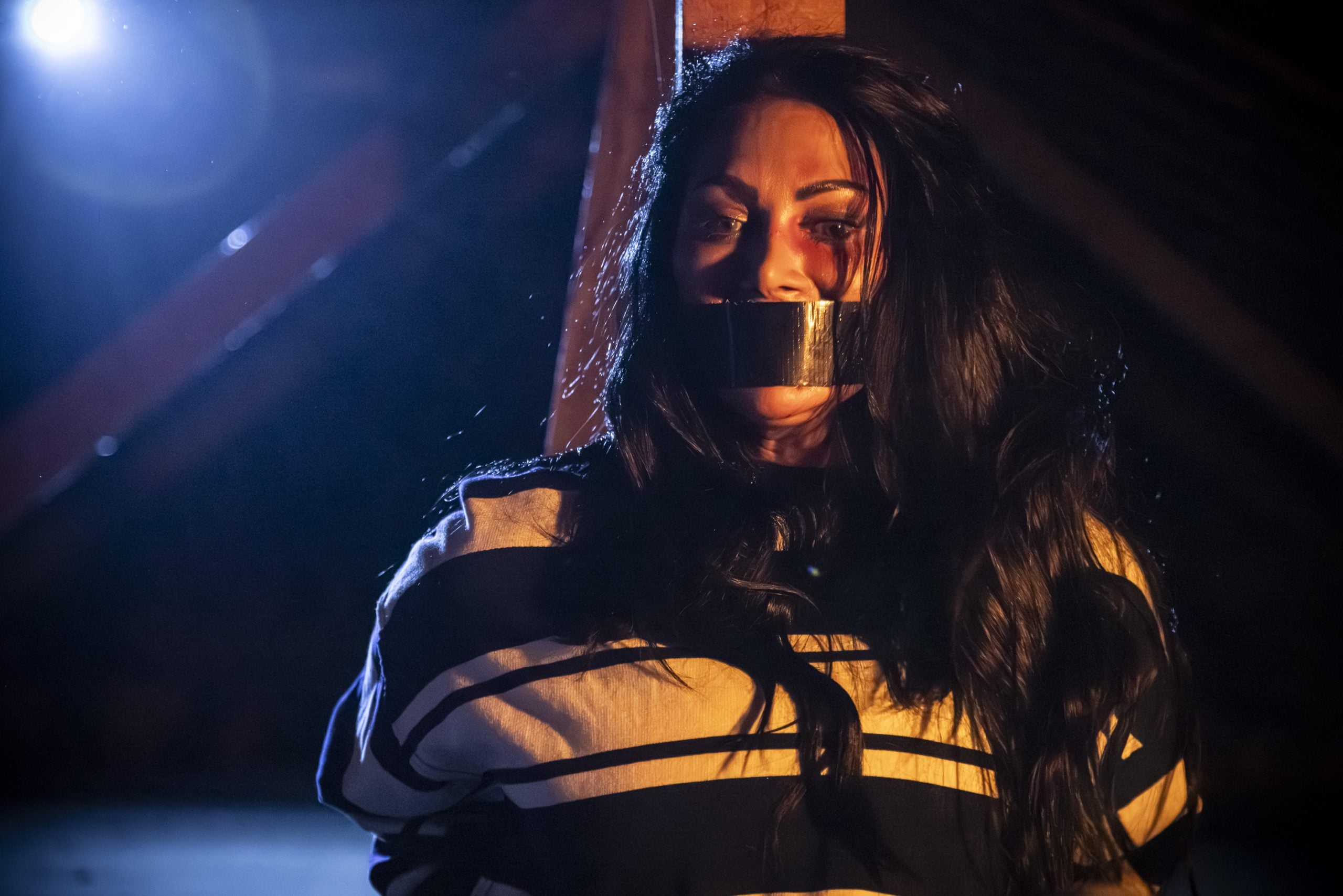 A woman with long dark hair, wearing a striped shirt, is sitting with her mouth covered by duct tape in a dimly lit space.