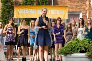 A group of young women stand outdoors in front of a “Welcome Back Theta Pi” banner, with one woman holding a microphone and speaking to the group.