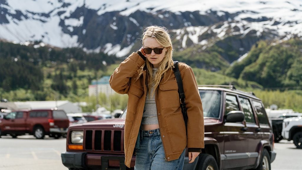A person in sunglasses and a brown jacket stands in a parking lot with a maroon Jeep and snow-capped mountains in the background.
