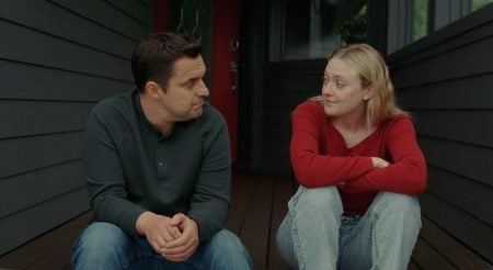 A man and a woman sit on a wooden porch outside a house, facing each other and talking. The man wears a dark shirt and the woman wears a red sweater and light jeans.