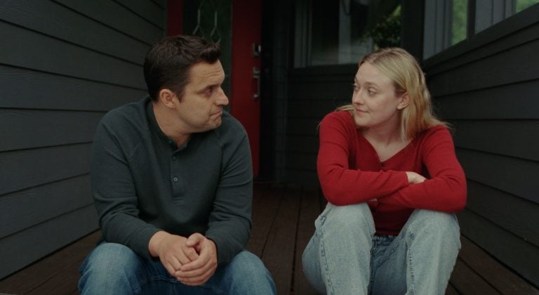 ‘The Sun Never Sets’ Review – A Raw And Honest Look At The Messiness Of Relationships And Old Wounds A man and a woman sit on a wooden porch outside a house, facing each other and talking. The man wears a dark shirt and the woman wears a red sweater and light jeans.