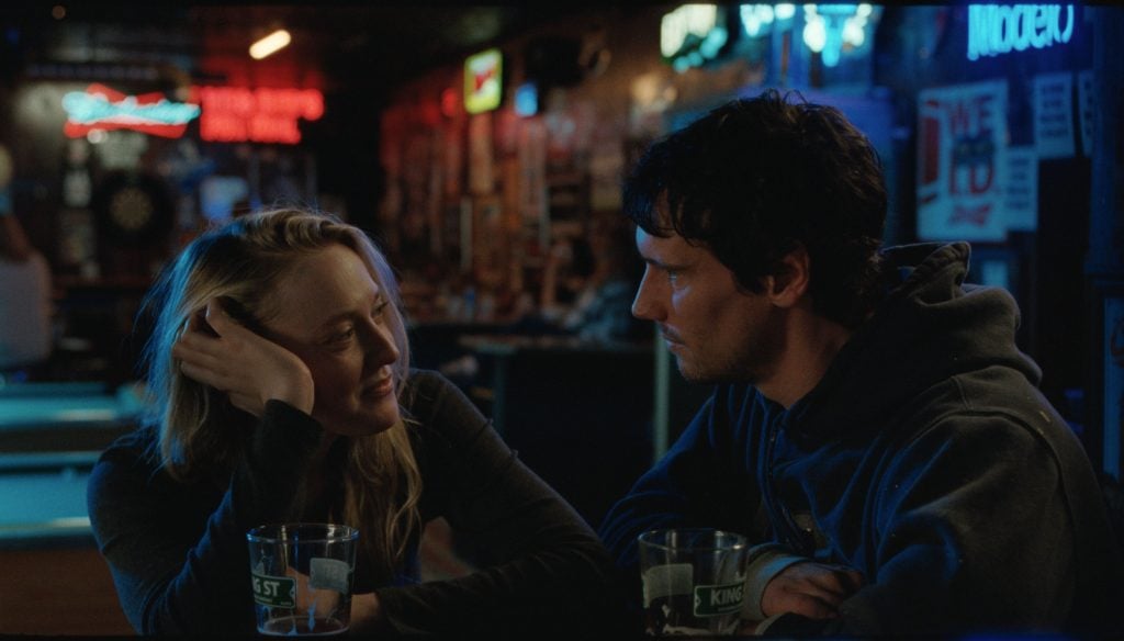 A woman and a man sit at a bar table with drinks, engaged in conversation, surrounded by neon lights and bar decor.