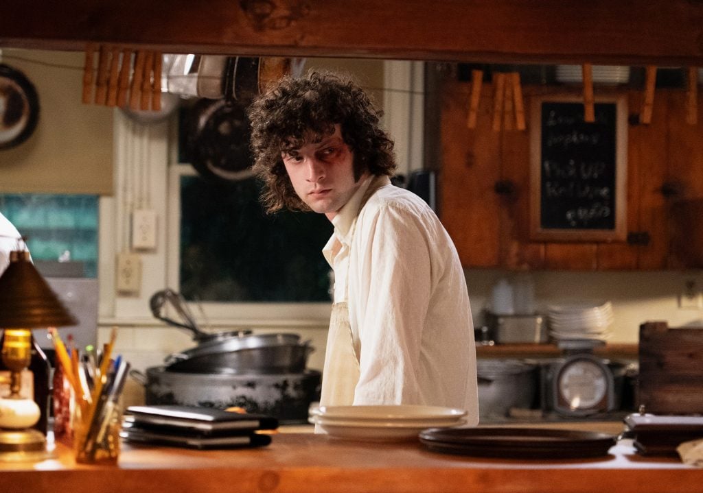 ‘Tony’ Review – A Beautiful Look At Growing Up And Finding Your Purpose A person with curly hair wearing a white shirt stands in a rustic kitchen, looking to the side, with dishes and kitchen utensils visible on the counter and shelves.