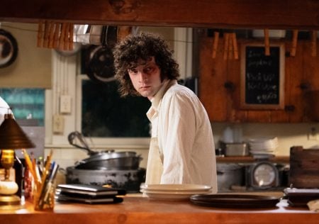 A person with curly hair wearing a white shirt stands in a rustic kitchen, looking to the side, with dishes and kitchen utensils visible on the counter and shelves.