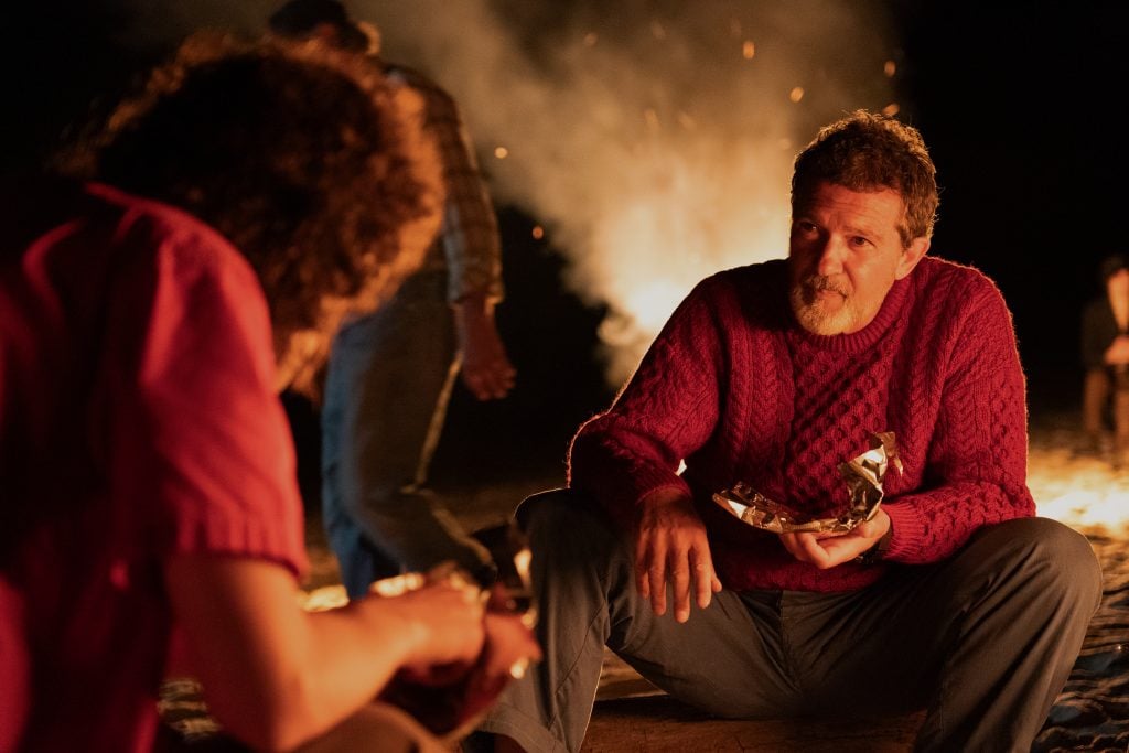 A man in a red sweater sits by a campfire at night, holding foil-wrapped food, talking to someone in a pink shirt.