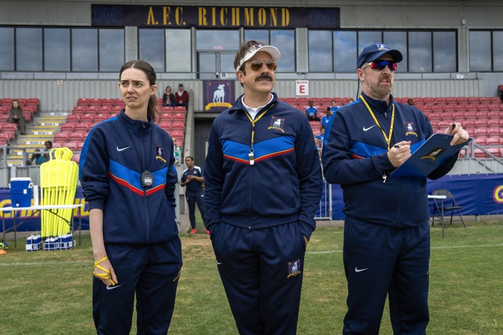 Three coaches in navy tracksuits stand on a soccer field, looking ahead. Stadium seats and “A.F.C. Richmond” sign are visible in the background.
