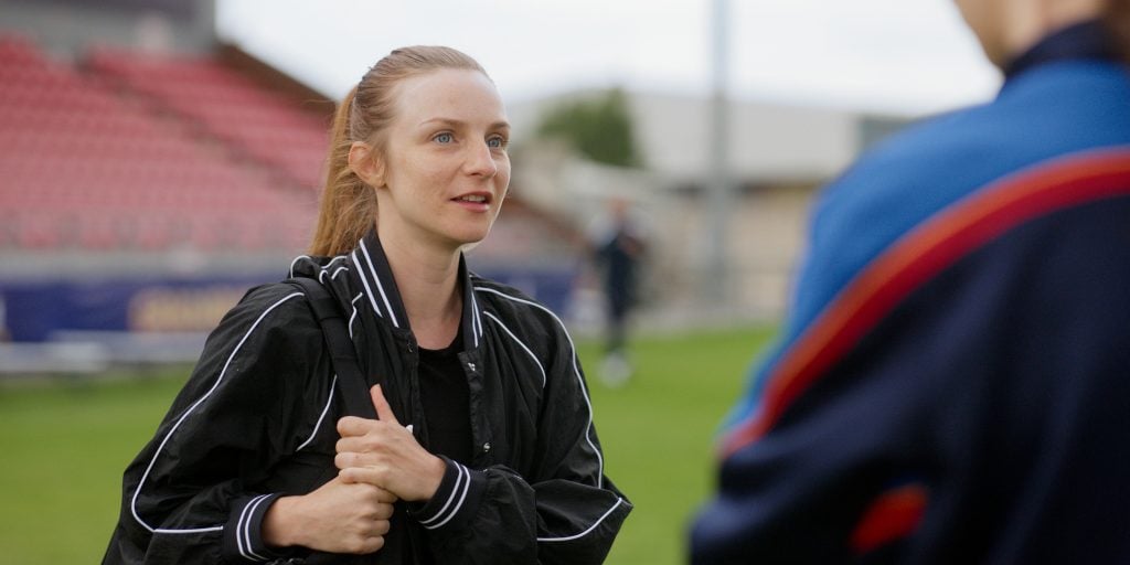 A woman in a black jacket stands outdoors on a sports field, holding a strap and talking to another person in a blue and red jacket.