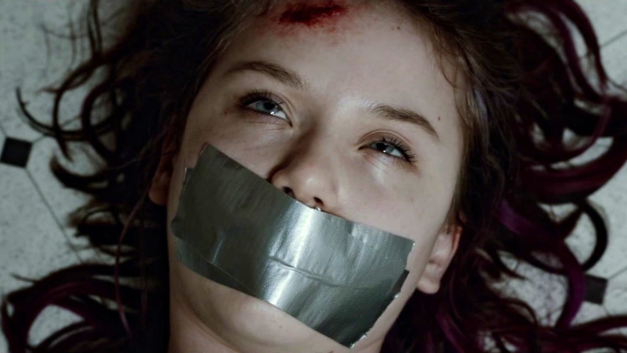 A person with a head wound lies on the floor with duct tape over their mouth, eyes open, and expression neutral.