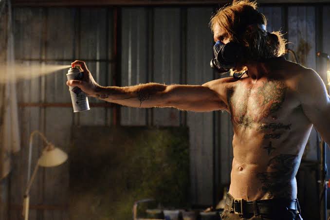 Shirtless person wearing a gas mask sprays paint from a can inside an industrial-looking space with metal walls.