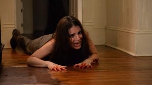 A woman with blood on her hands crawls on a wooden floor, looking distressed and scared, inside a dimly lit hallway.