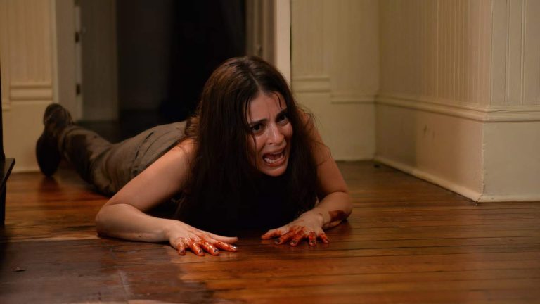 ‘The Devil’s Candy’ Second Sight 4K UHD Blu-Ray Review – Metal Horror Film Wrestles With A Lurking Evil A woman with blood on her hands crawls on a wooden floor, looking distressed and scared, inside a dimly lit hallway.