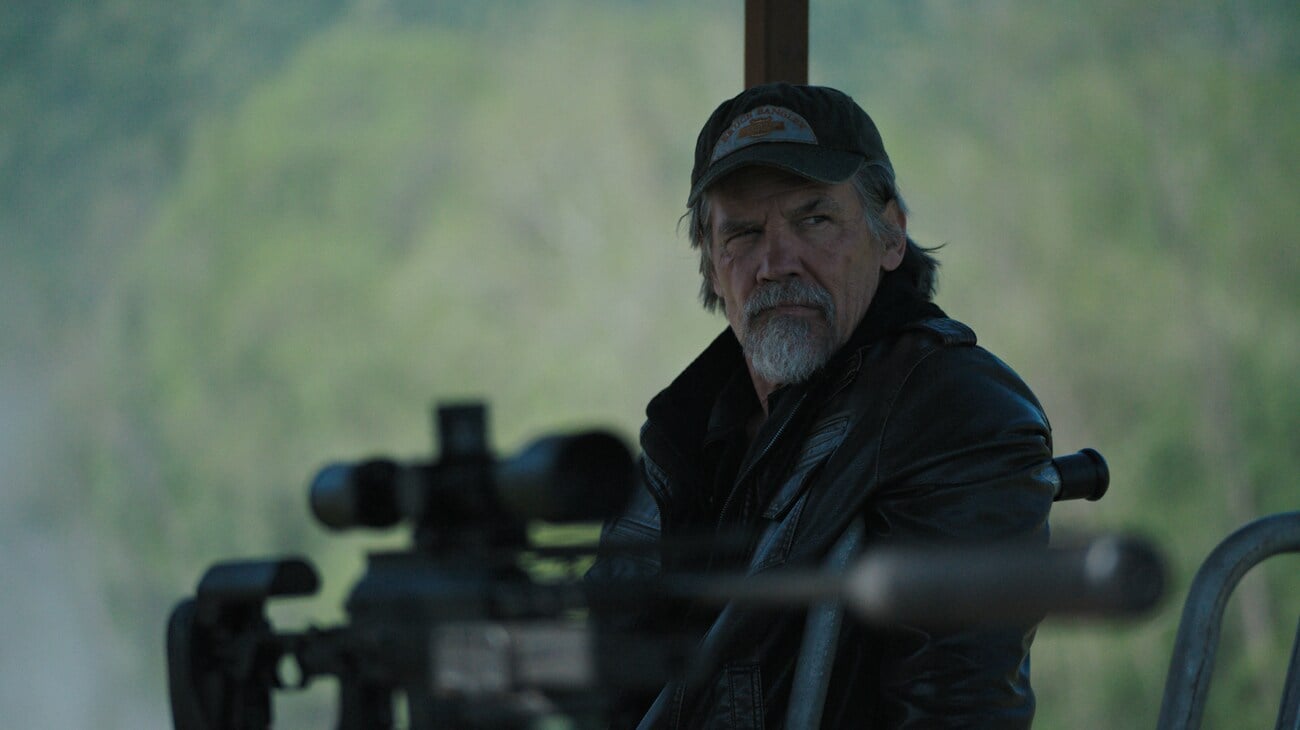 An older man with a gray beard and cap sits outdoors, wearing a black leather jacket, with a scoped rifle in the foreground.