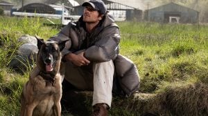 A man in a gray puffer jacket and cap sits on grass beside a brown dog. Farm buildings and green fields appear in the background.