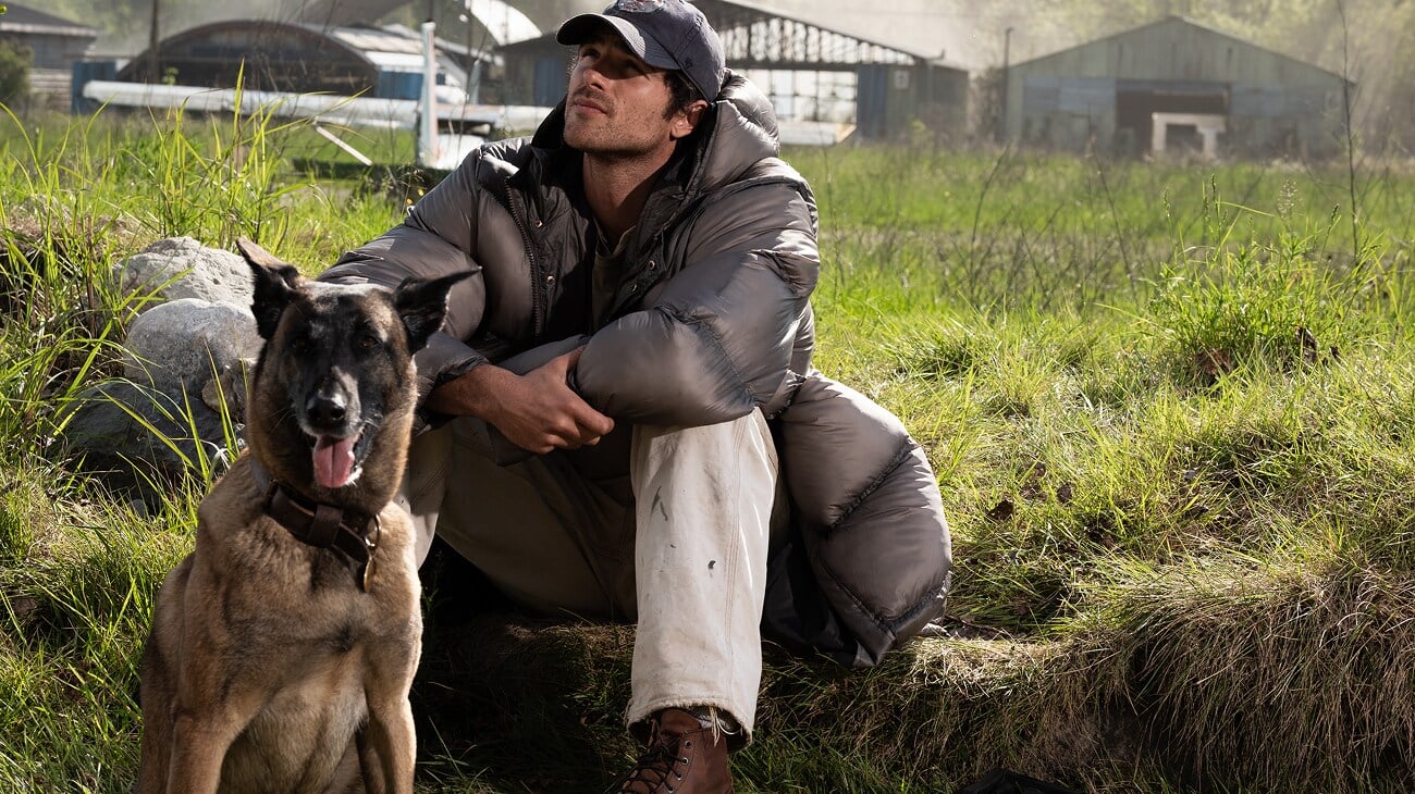 A man in a gray puffer jacket and cap sits on grass beside a brown dog. Farm buildings and green fields appear in the background.