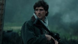 ‘The Dog Stars’ Review – Ridley Scott’s Post-Apocalyptic Picture Is An Admirably Meditative But Vapid Mixture Of Half-Baked Ideas A man in a green jacket and gray scarf holds a rifle outdoors, looking to his left with a concerned expression. The background is blurred and has a foggy, natural setting.
