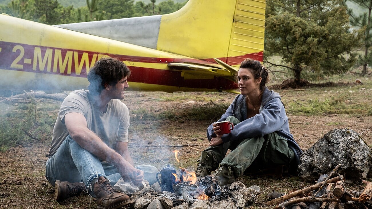 A man and woman sit by a campfire with mugs, next to a yellow and red plane that appears to have crash-landed in a wooded area.
