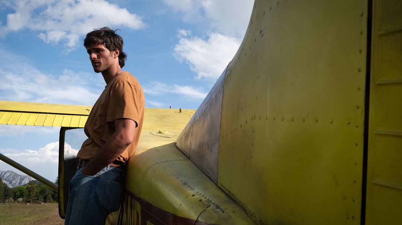 A man in a brown shirt and jeans stands with hands in pockets, leaning against the tail of a yellow airplane under a blue sky with clouds.