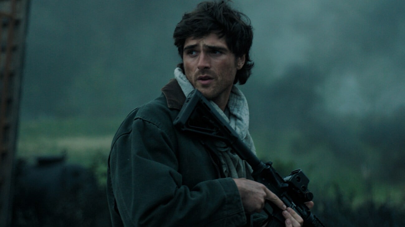 ‘The Dog Stars’ Review – Ridley Scott’s Post-Apocalyptic Picture Is An Admirably Meditative But Vapid Mixture Of Half-Baked Ideas A man in a green jacket and gray scarf holds a rifle outdoors, looking to his left with a concerned expression. The background is blurred and has a foggy, natural setting.