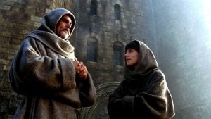 Two people in brown hooded robes stand outside a stone building, one older and bearded, the other younger, both facing each other and talking. Sunlight shines in from the upper right.