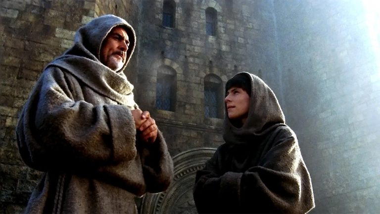 ‘The Name Of The Rose’ Second Sight 4K UHD Blu-Ray Review – Sean Connery Whodunnit Takes Us Back Centuries Two people in brown hooded robes stand outside a stone building, one older and bearded, the other younger, both facing each other and talking. Sunlight shines in from the upper right.
