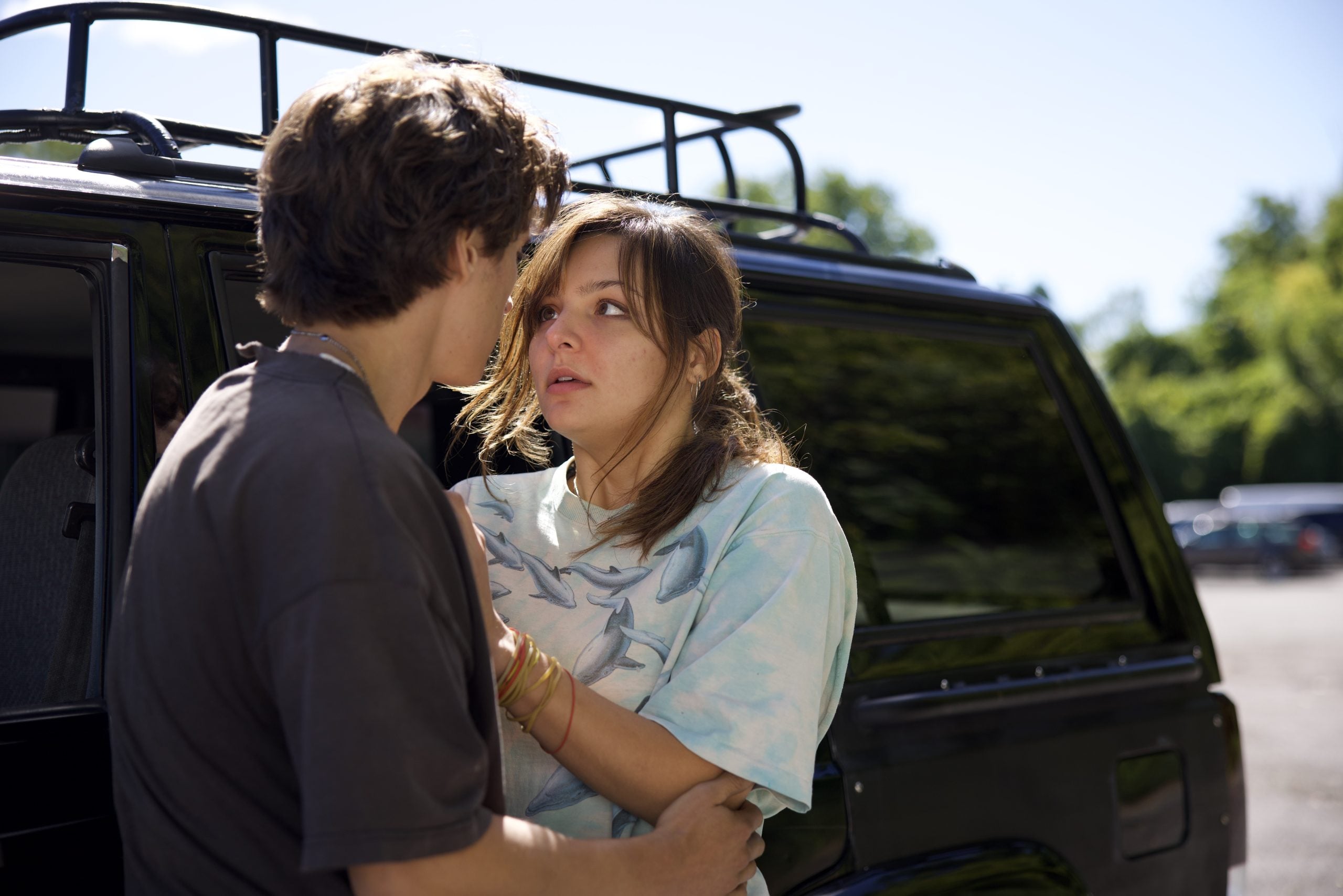 A young woman with a worried expression stands by a black SUV while a young man gently holds her arms; they appear to be having an intense conversation outdoors.