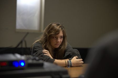 A young woman with long hair sits at a table, arms crossed, looking serious. A blurred figure is in the foreground, suggesting a conversation or interview setting.