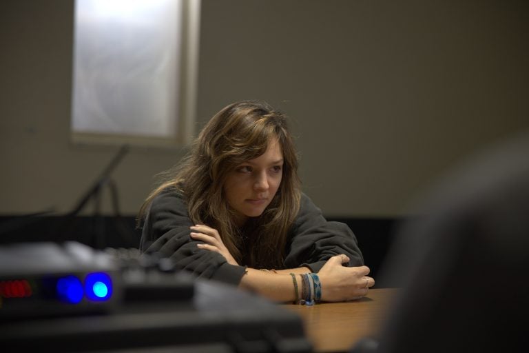 A young woman with long hair sits at a table, arms crossed, looking serious. A blurred figure is in the foreground, suggesting a conversation or interview setting.