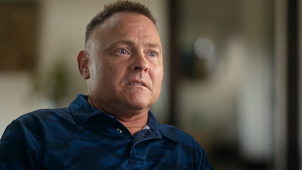 "Man with short hair in a blue shirt sits indoors, looking slightly off camera with a serious expression; background is softly blurred, suggesting a home or office environment."