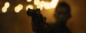 A person is pointing a handgun toward the camera in a dimly lit setting with blurred lights in the background.