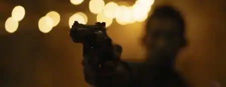 Zack Snyder Releases First Teaser For His Passion Project A person is pointing a handgun toward the camera in a dimly lit setting with blurred lights in the background.