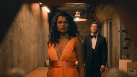 A woman in an orange dress stands in a dim hallway with a man in a suit behind her, both looking serious.