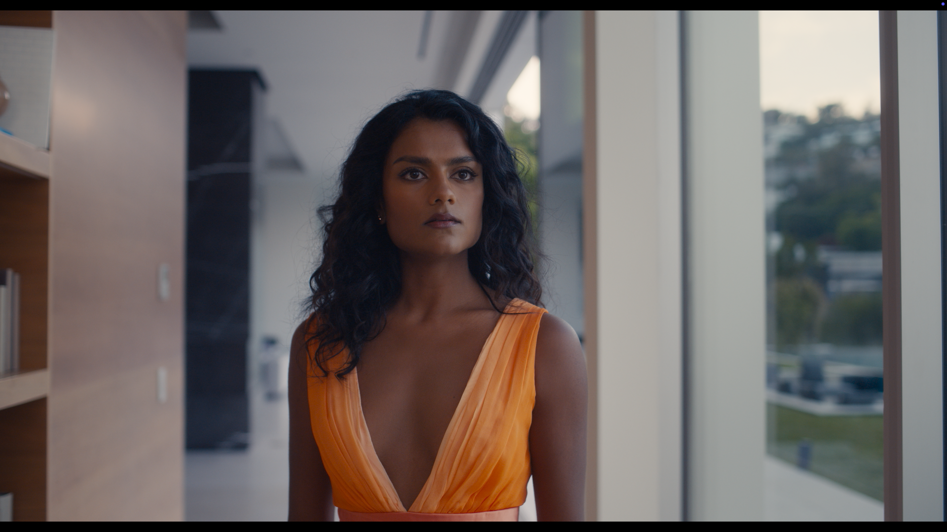 A woman with long dark hair in an orange dress stands indoors near large windows, looking serious.