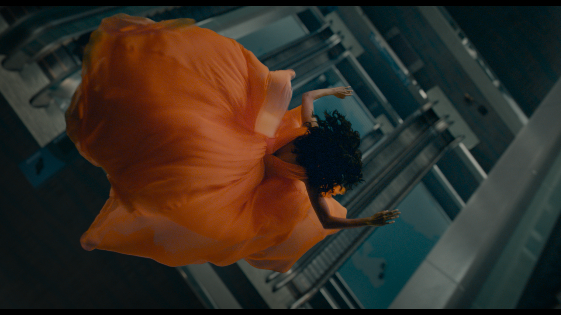 A person in an orange flowing dress is captured mid-air above escalators, viewed from above, arms outstretched.