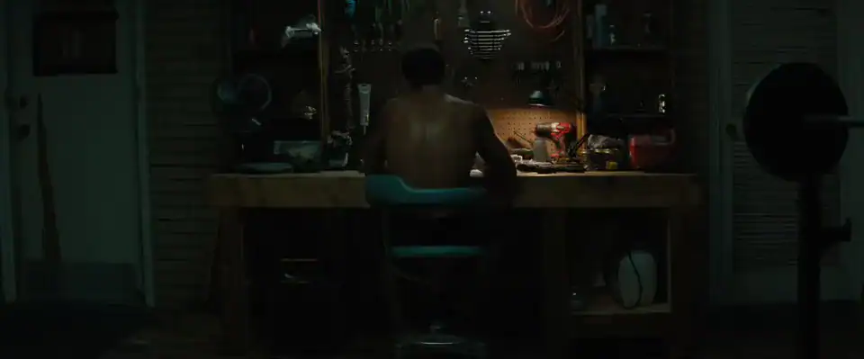 A shirtless person sits at a workbench in a dimly lit room, facing a wall with various tools, equipment, and supplies hanging above the workspace.