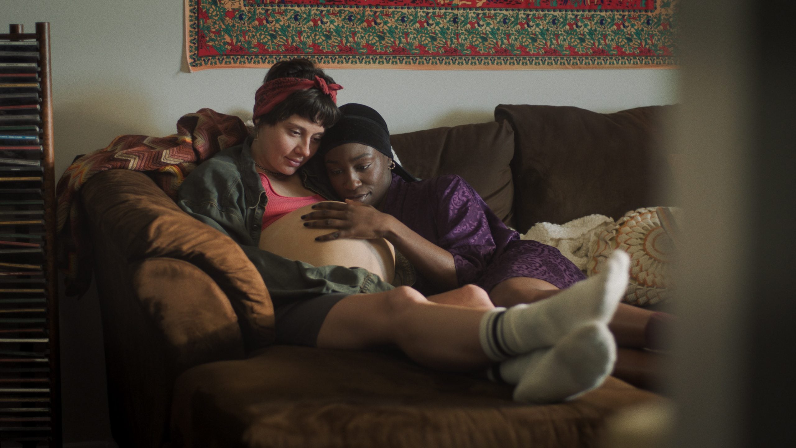 Two people sit closely on a brown couch; one is visibly pregnant while the other gently rests their head and hand on the pregnant person’s belly.