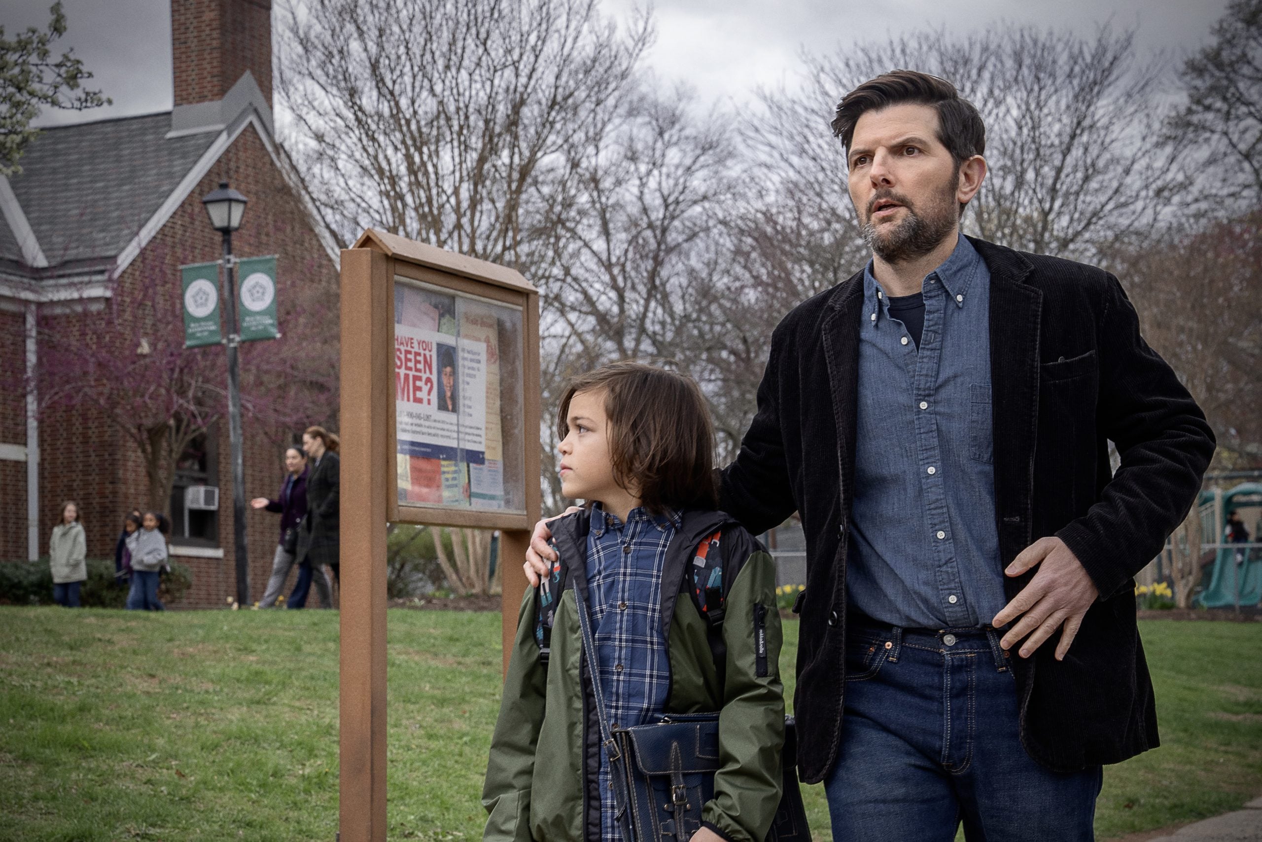 A man and a boy stand outside near a bulletin board with a “Have You Seen Me?” poster, with trees, people, and a brick building visible in the background.