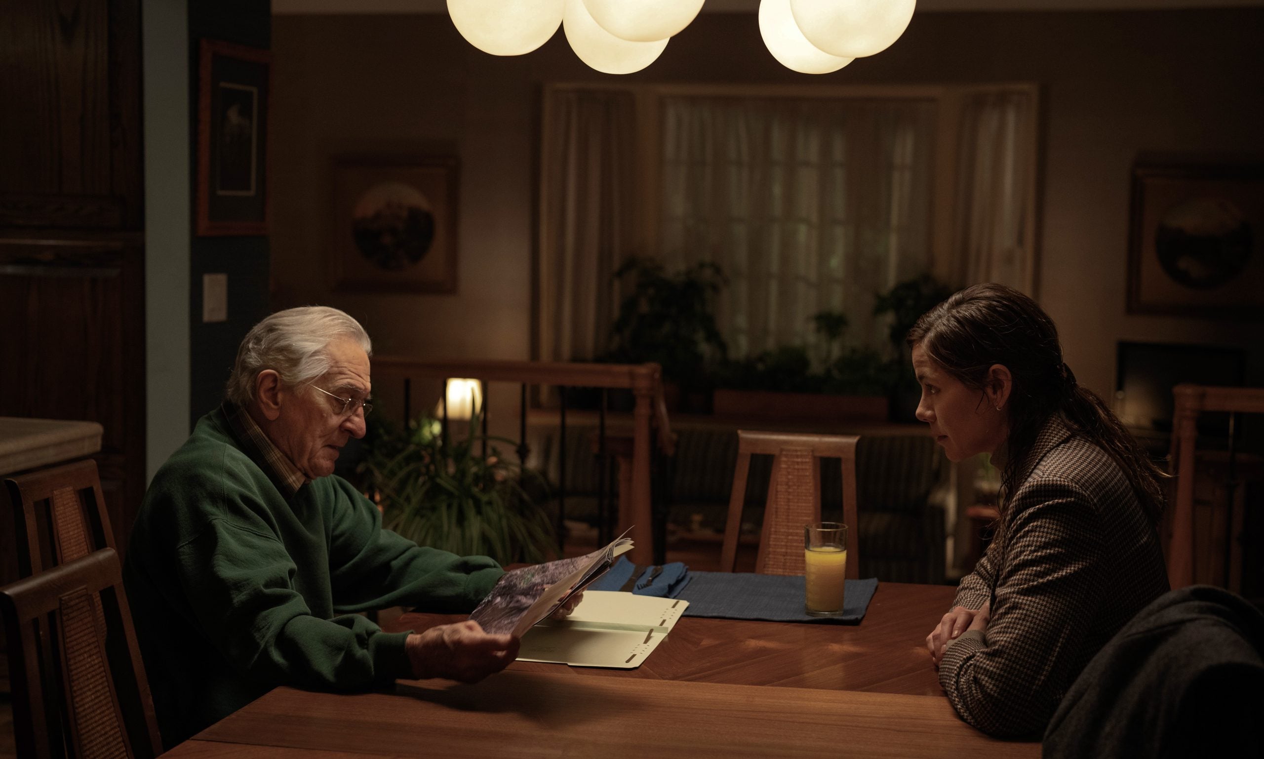 An older man and a younger woman sit across from each other at a dining table, talking. The man holds a document. The table has a glass of orange juice, folders, and blue placemats.