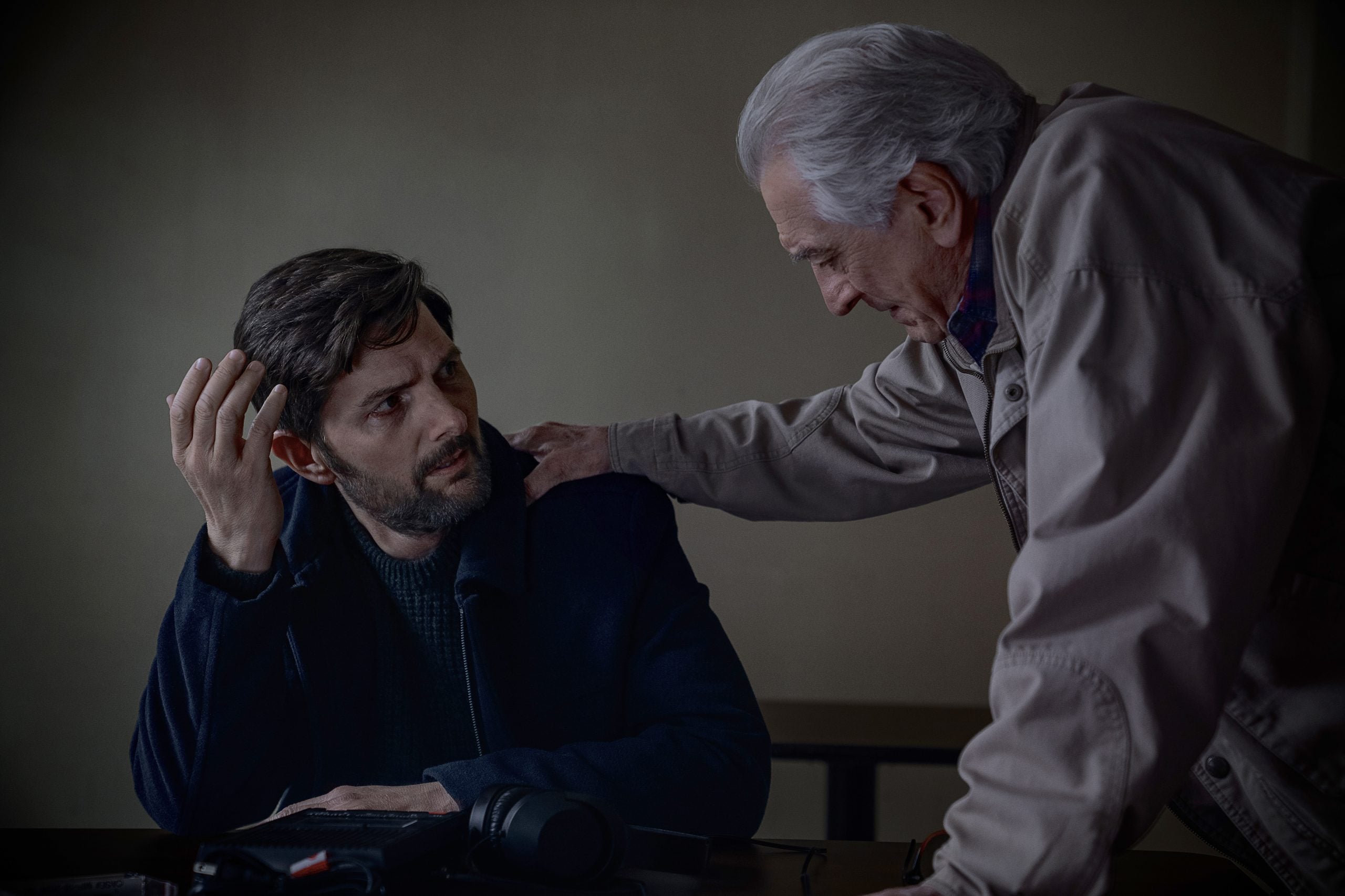 An older man stands beside a seated younger man, placing a hand on his shoulder while they have a serious conversation in a dimly lit room.