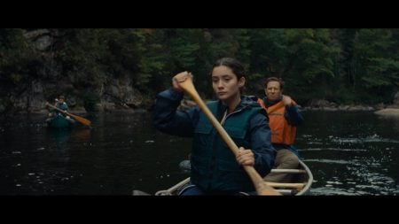 A woman and a man paddle a canoe on a calm river, surrounded by forested banks. Another canoe with two people is visible in the background.