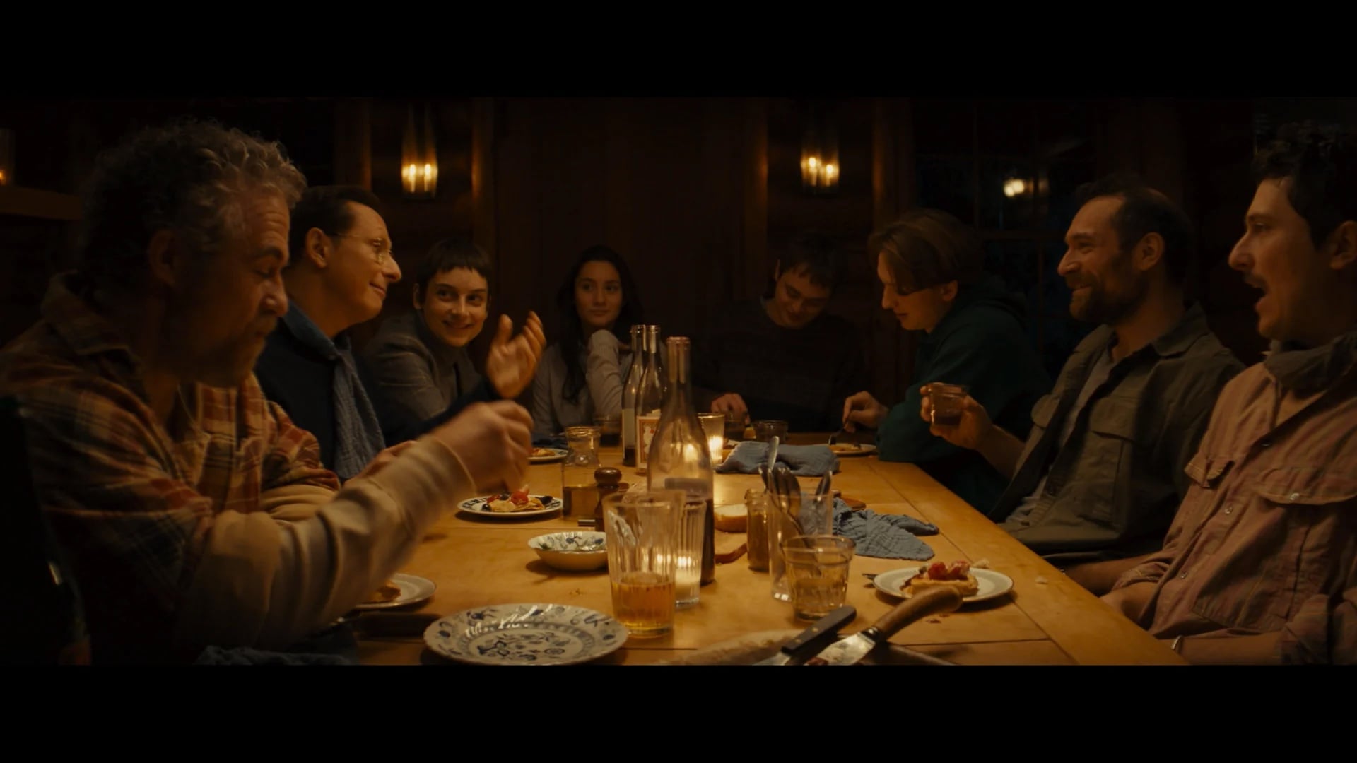 Eight people sit around a wooden dining table with plates, glasses, and bottles, engaged in conversation and sharing a meal in a warmly lit room.