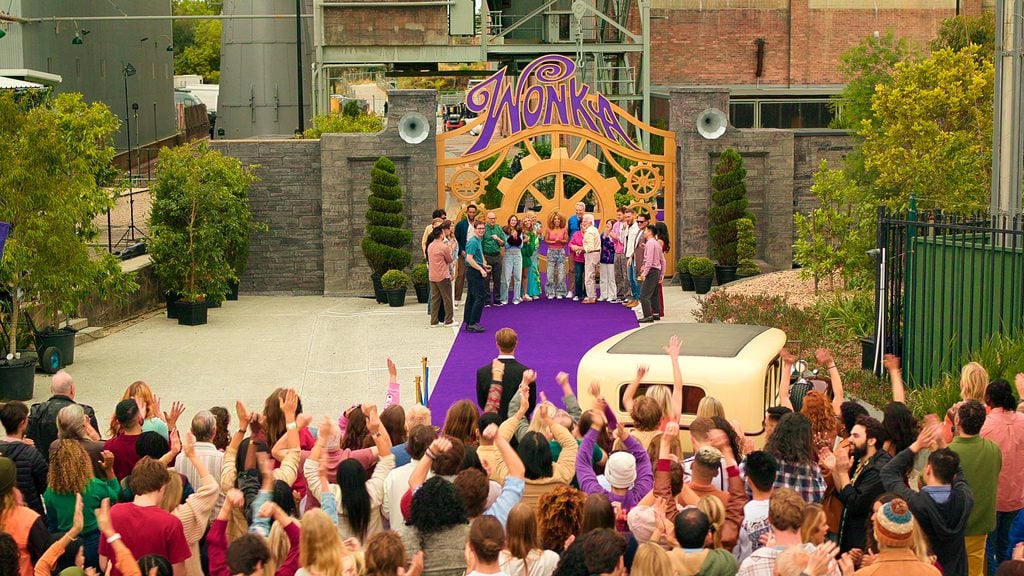 A crowd faces a stage with people standing under a large “Wonka” sign, set outdoors with a purple carpet and industrial background.