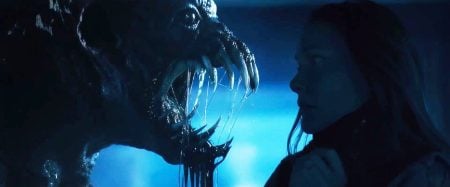 A woman faces a large, menacing creature with sharp teeth and drool dripping from its mouth in a dimly lit scene.