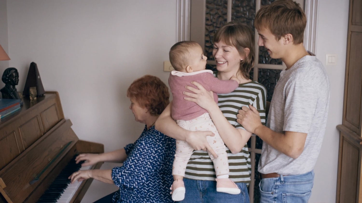 A woman plays piano while another woman holds a baby and smiles at a man standing beside her in a home setting.