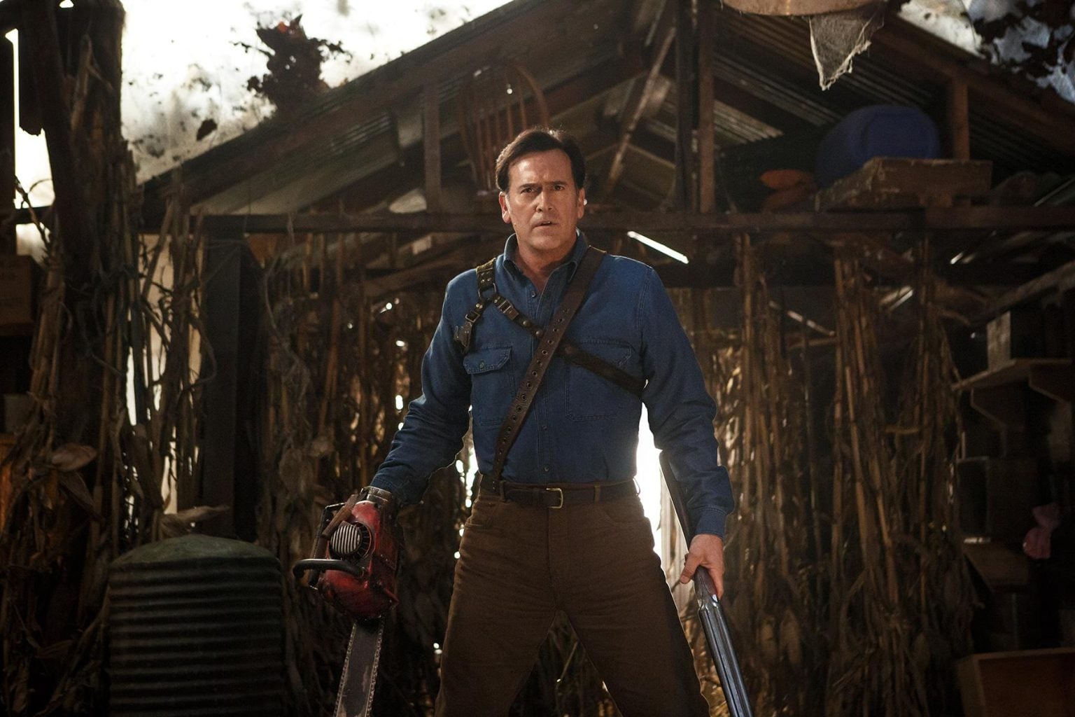 A man in a blue shirt stands in a rustic shed holding a chainsaw in one hand and a shotgun in the other, surrounded by corn stalks and wooden debris.