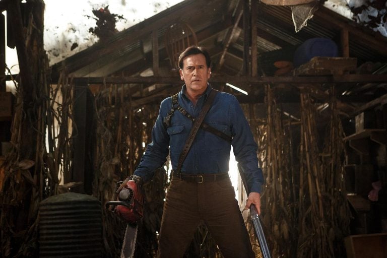 Bruce Campbell Has A Problem With Modern Evil Dead — And He Wants It Fixed A man in a blue shirt stands in a rustic shed holding a chainsaw in one hand and a shotgun in the other, surrounded by corn stalks and wooden debris.