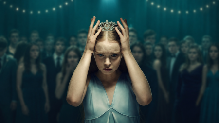 A young woman in a light blue dress stands at the center of a crowd, placing a tiara on her head, with string lights and formally dressed people in the background.