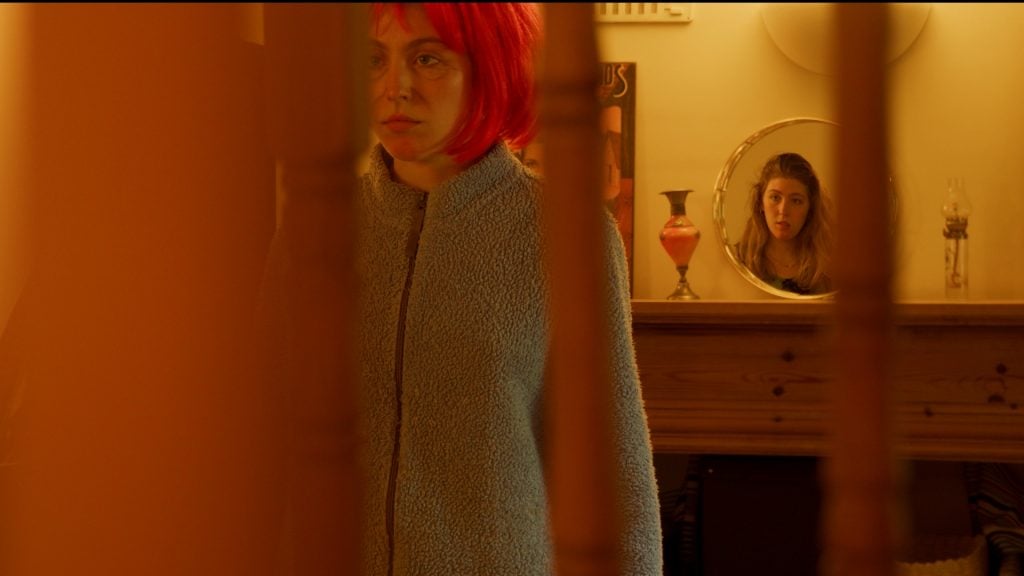 A woman in a red wig stands by wooden bars, while another woman’s reflection is visible in a round mirror on a mantelpiece in the background.