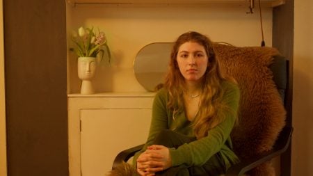 A woman with long hair sits on a fur-covered chair, hands clasped, in a room with a white vase of flowers and a mirror on a cabinet behind her.